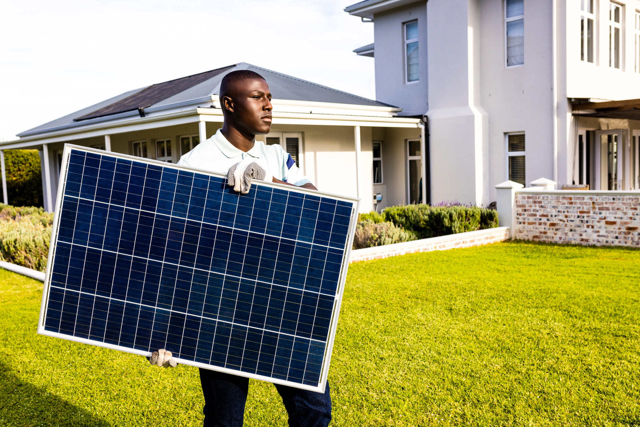 African american mid adult man carrying solar panel while walking in yard against house. Summer, unaltered, solar energy, electricity, green technology and sustainability concept.